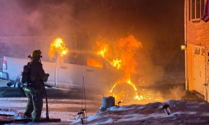 A firefighter sprays water on a white Ram truck that is on fire in a driveway at night.