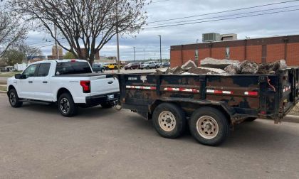 A white 2023 Ford F-150 Lightning pickup truck, likely a recent model year, is shown from the rear quarter view while connected to a dark-colored dual-axle trailer loaded with construction debris.