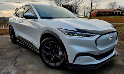 2023 Ford Mustang Mach-E in Star White Metallic parked on a suburban driveway, front three-quarter low angle view with a brick house and bare trees in the background.