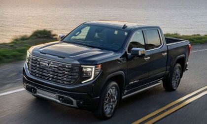A black 2023 GMC Sierra 1500 Denali truck drives on a coastal road at sunset, with ocean and greenery in the background. The scene conveys power and adventure.