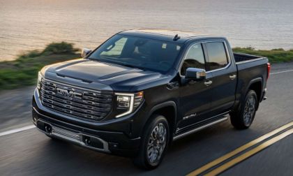 A black 2023 GMC Sierra 1500 Denali truck drives on a coastal road at sunset, with ocean and greenery in the background. The scene conveys power and adventure.