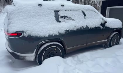 A dark-colored Rivian electric vehicle covered in heavy snow, showing the rear quarter view with illuminated taillights and snow-covered wheels in deep winter conditions.