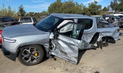 A severely damaged silver Rivian R1T, showing extensive side impact damage and crumpling along its passenger side while parked in what appears to be a salvage yard.