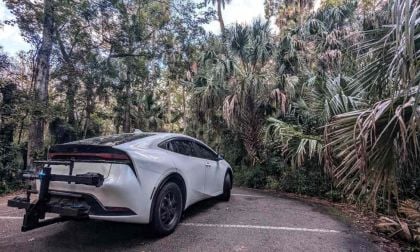 2023 white Toyota Prius hybrid with rear bike rack parked in a forest, rear three-quarter view.