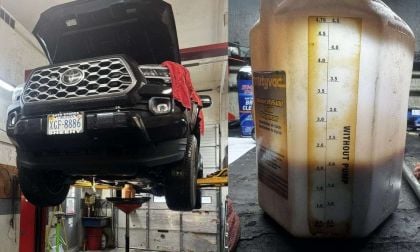 A 2023 Toyota Tacoma is elevated in a garage, with an oil catch pan below, alongside a fluid dispenser marked with measurements.