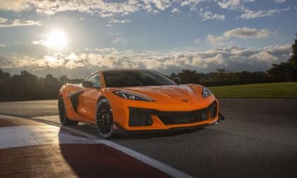 An orange Corvette stands on a racetrack, illuminated by sunlight, with dramatic clouds in the background and lush greenery in the distance.