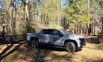 A silver 2024 GMC Sierra EV pickup truck shown from a front 3/4 angle, parked in a wooded area with tall pine trees in the background.