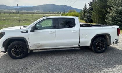 White 2024 GMC Sierra pickup truck with black wheels and off-road tires, photographed from side angle in mountainous rural setting with evergreen trees.