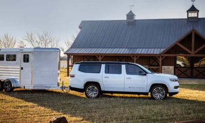A white 2024 Jeep Wagoneer towing a silver horse trailer near a rustic barn on grassy land, illuminated by warm sunlight.