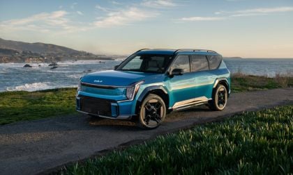 A teal blue 2024 Kia EV9 electric SUV is shown from a front three-quarter angle, parked on a coastal overlook with dramatic ocean waves and mountains in the background.