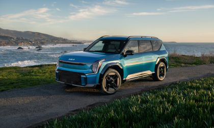 A teal blue 2024 Kia EV9 electric SUV is shown from a front three-quarter angle, parked on a coastal overlook with dramatic ocean waves and mountains in the background.