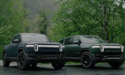 Two 2023 Rivian R1S And Rivian R1T electric pickup trucks in forest green, photographed from front 3/4 view, featuring distinctive LED light bars and black wheels, parked in wooded setting.