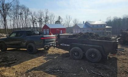 Pickup truck towing a trailer filled with soil, near red and white buildings, surrounded by trees.