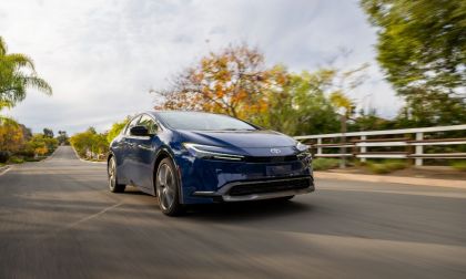 A navy blue 2024 Toyota Prius shown from a front three-quarter angle, driving on a tree-lined suburban road with fall foliage in the background.
