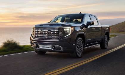 Black 2024 GMC Sierra 1500 driving on a coastal road at sunset, shown from the front three-quarter angle.