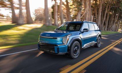 Blue Kia EV9 electric SUV driving on a forest-lined road with motion blur highlighting smooth performance.