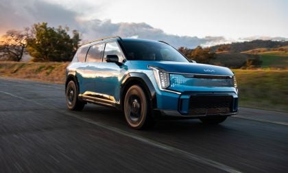 Blue 2024 Kia EV9 electric SUV parked on a mountain road with green hills and dramatic clouds in the background.