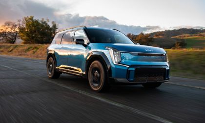 Blue 2024 Kia EV9 electric SUV parked on a mountain road with green hills and dramatic clouds in the background.