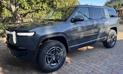 2024 Rivian R1S in dark gray parked on a brick driveway with a large tree and suburban house in the background.