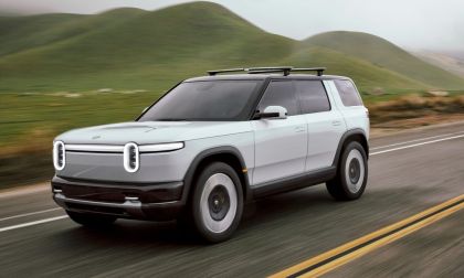 Silver Rivian R2 electric SUV driving on a paved road through rolling green hills with motion blur emphasizing speed.