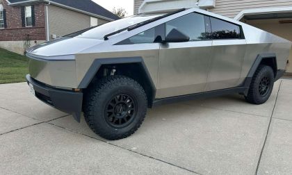 Side view of a Tesla Cybertruck in metallic silver, showcasing its angular design and black off-road wheels with aggressive all-terrain tires parked on a concrete driveway.