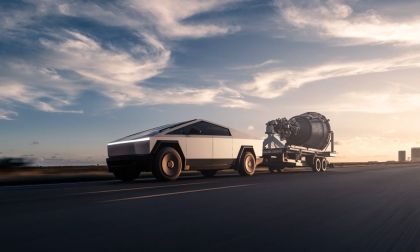 Tesla Cybertruck towing a large industrial turbine on a trailer while driving on an open highway at sunset.