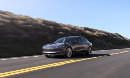 2024 Tesla Model 3 in Midnight Silver Metallic driving on a coastal highway, front three-quarter view with hillside and blue sky background