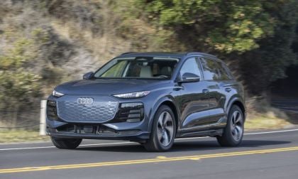 2024 Audi Q6 e-tron in metallic gray, front three-quarter view, driving on winding mountain road with distinctive LED headlights and large octagonal grille.