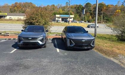 2026 Cadillac Optiq electric SUV in metallic gray, parked front-end view in a retail parking lot with fall foliage and Dollar General store visible in background.