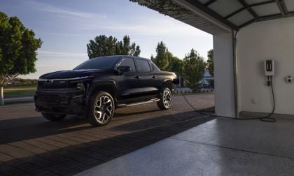 Electric pickup truck charging in a driveway, connected to a wall-mounted charger in a garage.