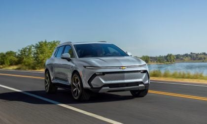 Silver 2025 Chevy Equinox EV driving on lakeside highway, sleek electric SUV with modern grille design