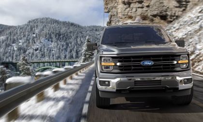 A silver 2025 Ford F-Series pickup truck shown from the front quarter angle, driving on a mountain road with snow-covered peaks in the background.