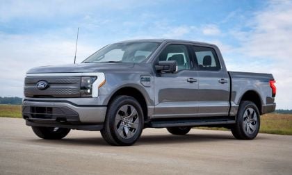 2022 Ford F-150 Lightning electric pickup truck in silver, three-quarter front view, featuring distinctive LED light bar and closed grille, parked on concrete with rural landscape background.