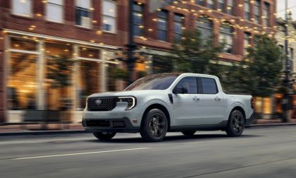 A white 2023 Ford Maverick pickup truck shown from a front three-quarter angle, driving on a city street with motion blur and brick buildings in the background.