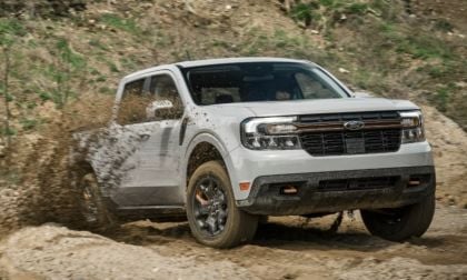 White pickup truck driving through muddy terrain, splashing mud around, with grassy background.