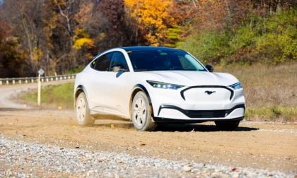 White Ford Mustang Mach-E electric SUV driving on gravel road with autumn forest background