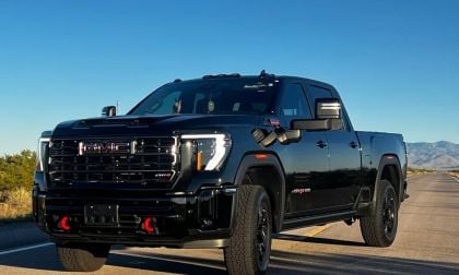 Black 2024 GMC Sierra HD pickup truck photographed from front quarter angle, featuring bold chrome grille, lifted suspension, and off-road tires, parked on desert highway with mountains behind.