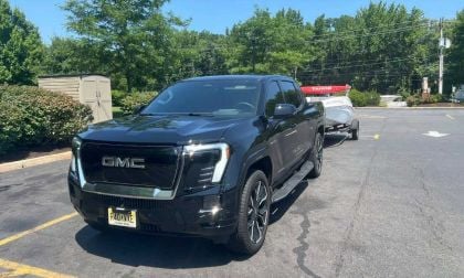 Dark blue 2025 GMC Sierra EV pickup truck parked in suburban lot with trees and utility pole in background, angled front three-quarter view showcasing bold grille and unique design.