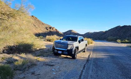 2025 GMC Sierra EV electric pickup truck parked on a desert roadside, showing front three-quarter view with rugged terrain and mountains in the background