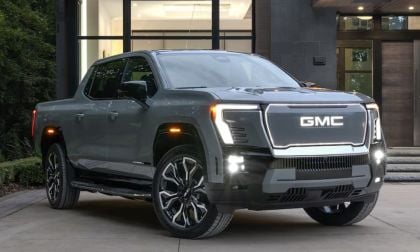 Dark gray 2025 GMC Sierra EV Denali pickup truck parked in front of modern glass and stone building with LED headlights on