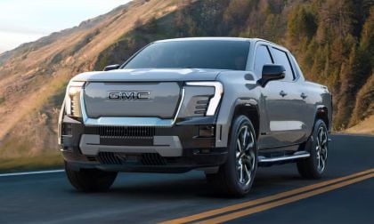 Silver GMC Sierra EV pickup truck driving on mountain road with pine forest backdrop and winding highway