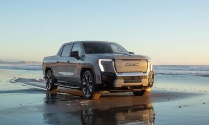 2025 GMC Sierra EV Denali pickup truck in silver parked on a reflective beach at sunset, showcasing its futuristic LED headlights and bold electric truck design.