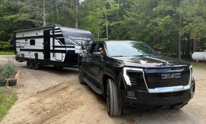 Black GMC Sierra EV towing white Transcend travel trailer through wooded campground with dense forest background