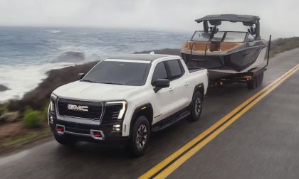 White GMC Sierra AT4X pickup truck, front three-quarter view, towing boat on coastal highway with ocean backdrop.