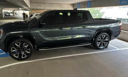 A silver 2025 GMC Sierra EV pickup truck shown from a high three-quarter front angle, featuring black accents, a dark tinted panoramic roof, and distinctive LED lighting elements.