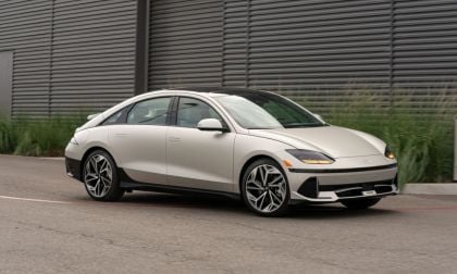A white sedan parked on a street beside a corrugated metal building and greenery.