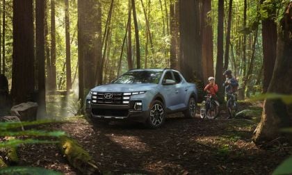 A silver Hyundai pickup truck is parked in a sunlit forest, surrounded by tall trees. Two cyclists in gear stand nearby, evoking adventure and exploration.