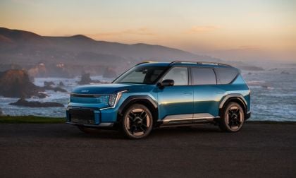 Blue 2025 Kia EV9 electric SUV parked near an ocean cliff at sunset, front three-quarter view.
