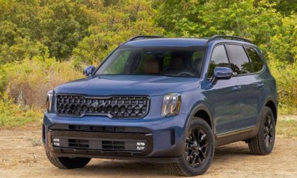 A blue 2025 Kia Telluride SUV shown from a front three-quarter angle, featuring its distinctive black grille and rugged off-road styling against a natural wooded background.