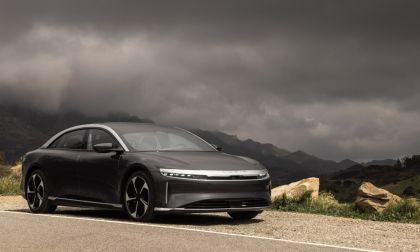 A black Lucid Air electric luxury sedan shown from a front three-quarter angle, parked on a mountain road with dramatic cloudy skies in the background.